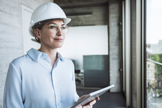 Thoughtful engineer wearing hardhat standing with tablet PC in office