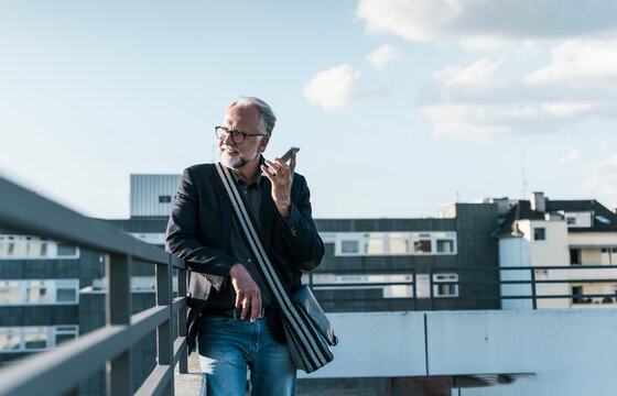 Happy businessman talking through speaker phone standing near railing on terrace