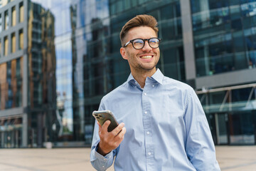Elegant gentleman in a blue shirt smiling with a smartphone in hand, against a modern glass...