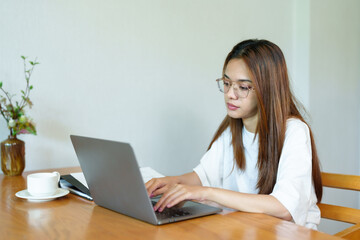 Asian woman wearing glasses sitting at wooden table working on laptop, white cup and saucer beside, casual attire, indoor home office setting, daytime. Bright natural light