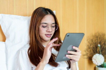 Fototapeta premium Beautiful Asian woman with glasses sitting in bed, holding a tablet, watching a movie. Wearing casual white t-shirt, relaxed posture, Indoor bedroom setting, comfortable and cozy atmosphere.