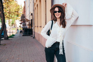 Portrait of young beautiful smiling hipster woman in trendy summer white blouse and jeans. Carefree woman posing in the street in sunny day. Positive model outdoors. Cheerful and happy, in sunglasses