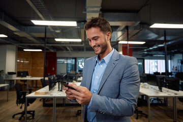 Fototapeta premium Happy young caucasian man in businesswear using mobile phone in office