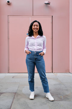 Smiling businesswoman standing with hands in pockets in front of pink wall