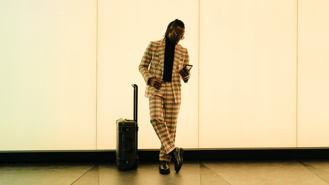 Businessman using smart phone standing by luggage near illuminated wall