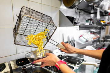 Close-up of hands lifting a basket of freshly fried French fries from a deep fryer in a commercial...
