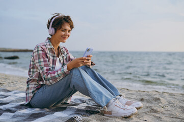 Full body young woman wear shirt casual clothes sits listen to music in headphones use mobile cell...