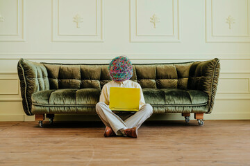 Businessman with mask of chaotic thoughts sitting on floor and using laptop