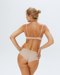 Naklejka premium Rear view of blonde woman in beige lingerie posing holding hands on hips against white studio background. Concept of beauty and female health, lifting, cosmetology procedures, body care, antiaging. Ad