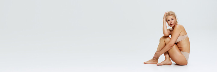 Obraz premium Banner. Posed in nude lingerie, blonde woman sits with one hand on her head against white studio background with negative space. Concept of beauty, cosmetology procedures, body care, antiaging. Ad