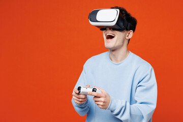 Young man he wearing blue sweatshirt casual clothes hold in hand play pc game with joystick console watching in vr headset pc gadget isolated on plain red orange color background. Lifestyle concept.