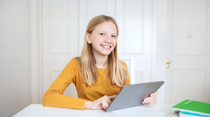 Fototapeta premium A teen girl with long blonde hair sits at a table, smiling as she uses a tablet computer. The table is white, and the background is a white wall with a closed door.