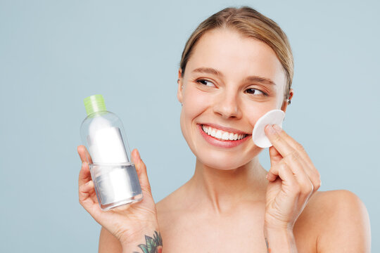 Half naked young woman with nude make up cleaning face using cotton pad and cleansing micellar product look aside isolated on plain blue background. Skin care healthcare cosmetic procedures concept.