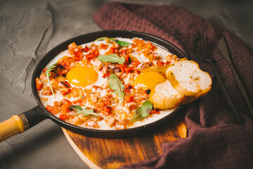 Shakshuka, scrambled eggs with tomatoes, capsicum, bacon and onions in a cast iron pan with toasted bread on a gray background with a knife and fork