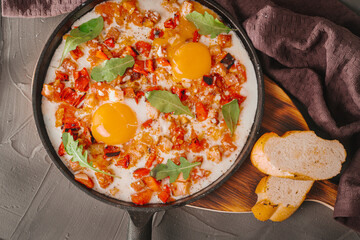 Scrambled eggs with tomatoes, peppers, shallots and bacon in a cast iron skillet with slices of bread. Delicious, nutritious and healthy breakfast Gray background, burgundy napkin. High quality photo