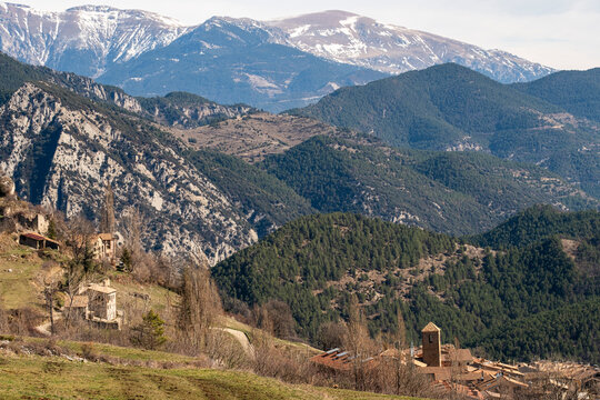 A beautiful mountain valley featuring scattered rustic houses and lush green forests, all set against a backdrop of towering snow-capped peaks and vast blue skies in Pedraforca in Catalonia