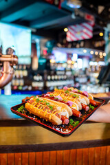 Close-up of hot dogs with various toppings on a plate, held in a bar or restaurant setting. Blurred background includes bottles. Perfect for food, dining, and American culture themes.