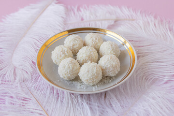 White Chocolate Candy With Coconut Topping On White Feather and pink background, sweet balls on plate flat lay. Tasty gift for any celebration and anniversary