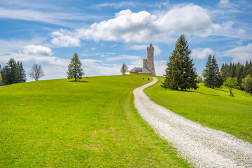 A winding gravel path leads up to the Dalimil Lookout Tower in Czechia, surrounded by lush green fields and towering trees. The sky is a bright blue with fluffy white clouds.