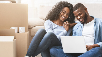 Smiling african couple sitting on floor at new apartment, choosing new furniture online, using laptop, copy space