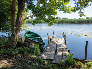 A peaceful riverside view featuring a green wooden boat tied to an old wooden dock under the shade of trees, with calm water and lush vegetation in the background