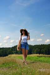 Obraz premium beautiful dark-haired woman walking casually across a field, wearing a sun hat and jeans shorts