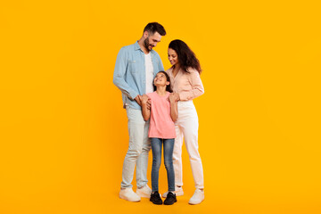 A happy family stands together in front of a yellow background, smiling at each other. They all...