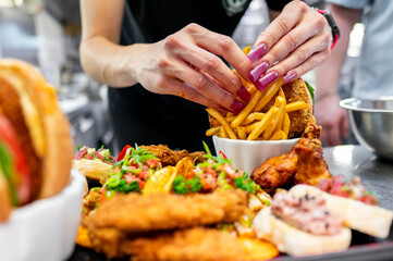 Close-up of hands with purple nails serving French fries in a fast-food restaurant. Blurred burgers and fried chicken in the background. 