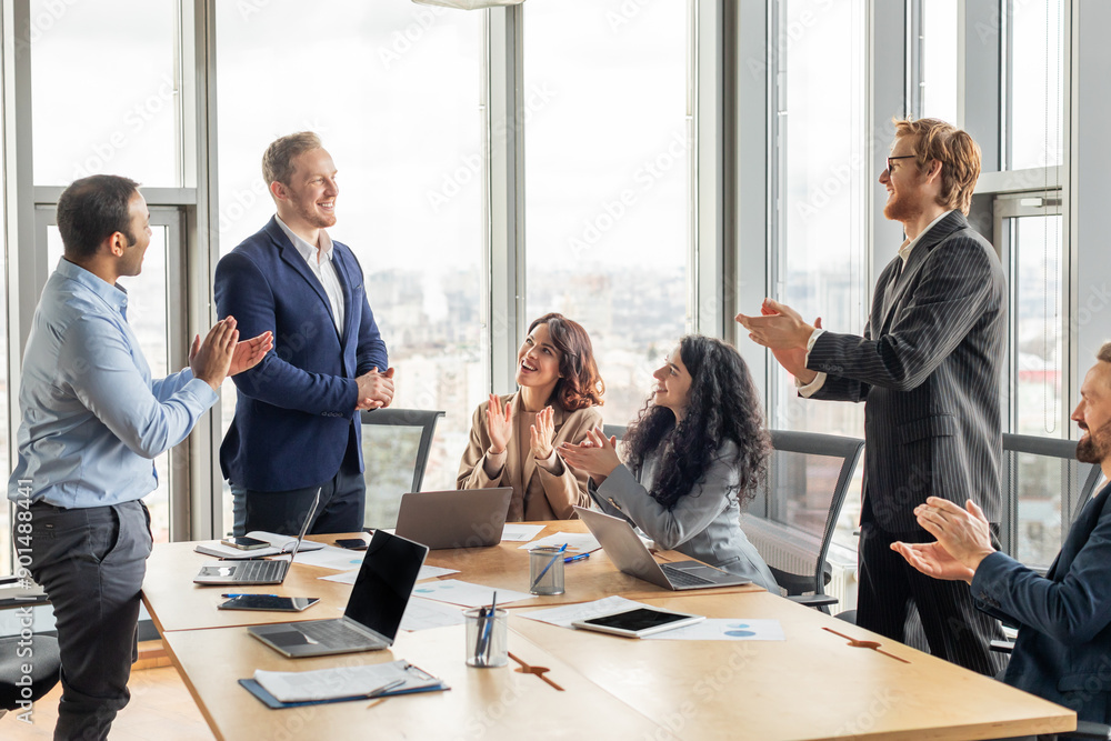 Aufkleber A business team is gathered around a conference table in a modern office with large windows. They are all clapping and smiling as they applaud a colleague who is standing in front of them.