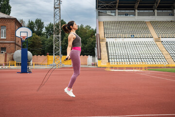 A young sporty woman doing her morning workout, skipping rope on the stadium, outdoors, side view