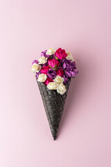Black ice cream cone with colorful flowers on pastel pink background. Minimal summer composition.