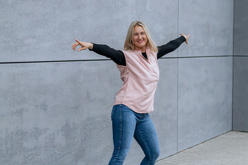 Happy positive dancer curling in dance. Woman with blond hair dancing on urban street, on gray wall...