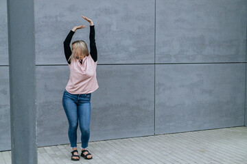 Happy positive dancer curling in dance. Woman with blond hair dancing on urban street, on gray wall...