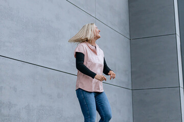 Happy positive dancer curling in dance. Woman with blond hair dancing on urban street, on gray wall...