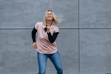 Happy positive dancer curling in dance. Woman with blond hair dancing on urban street, on gray wall...