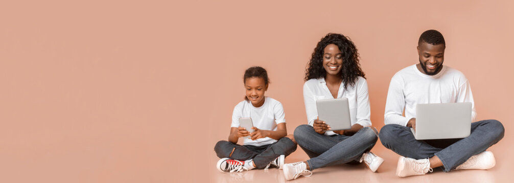 Modern family. Happy african american father, mother and daughter using different gadgets while sitting on floor over yellow studio background, panorama