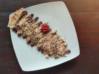 Sweet dessert on a white plate on a wooden table.  Strawberries, peanut flakes and chocolate on a tortilla.  Peanuts sweet tortillas.  Soft focus.  Negative space.  Semi flatlay.