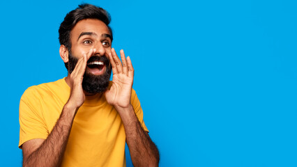 Indian young man with a beard is standing in front of a blue background. He is cupping his hand...