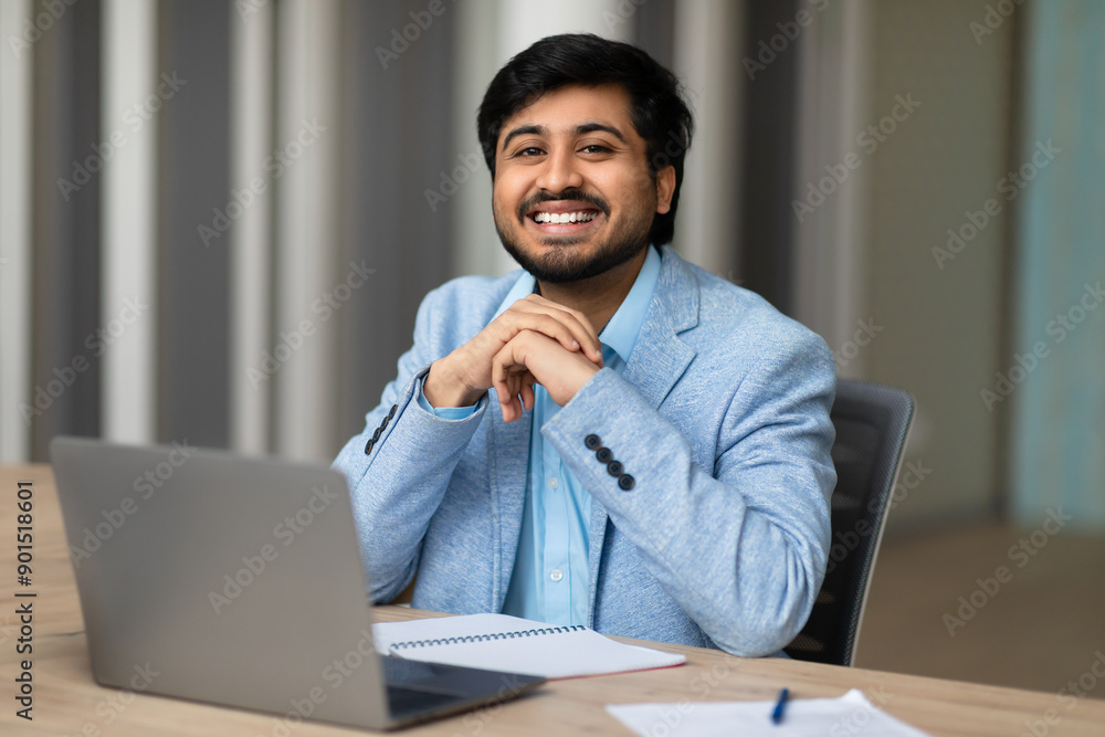 Aufkleber a young indian man in a light blue blazer sits at a desk in a modern office. he is looking directly 