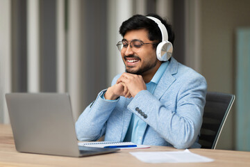 Indian man wearing headphones and glasses sits at a desk and smiles while looking at a laptop screen. He is likely participating in a video call with someone else. A notebook and pen are on the desk