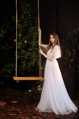 A caucasian woman in an elegant white lace dress sits on a wooden swing amidst dark foliage.