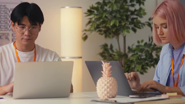 Pink-haired woman designer and her Asian colleague working on laptops in office