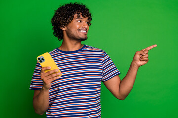 Portrait of young curly haired guy in striped t shirt using smartphone direct finger mockup interested isolated on green color background