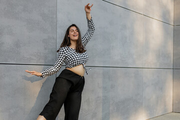 Happy positive dancer curling in dance. Woman with long dark dancing on urban street, on gray wall...