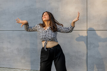 Happy positive dancer curling in dance. Woman with long dark dancing on urban street, on gray wall...