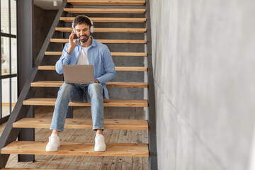 Fototapeta premium A man sits on a wooden staircase with headphones on, working on a laptop while smiling. He is wearing blue jeans and a light blue button-down shirt, copy space