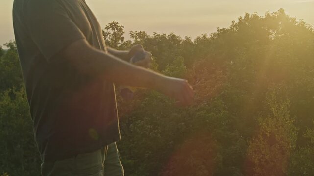 During a picturesque sunset on a mountain, a man sprays mosquito repellent. The vibrant colors of the sky and peaceful setting enhance the scene.