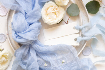 Cards near light blue fabric and cream flowers on plate top view, copy space, wedding mockup
