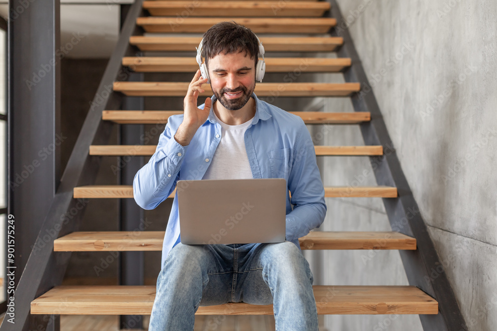 Poster a man sits on a staircase with a laptop in his lap. he is wearing headphones and adjusting them whil