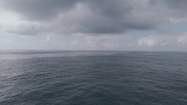 Flying over the open ocean, a drone captures slow-motion waves. Cloudy skies with some sunlight breaking through. The horizon unites sky and sea.